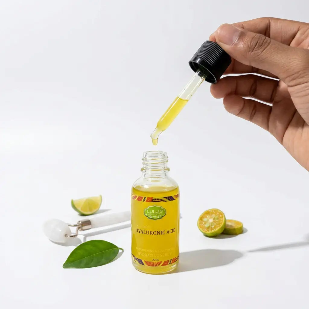 Hand holding a glass dropper with golden serum above a bottle of Lulu's Holistics Hyaluronic Acid Hydrating and Lightening Serum next to a face roller and fresh limes