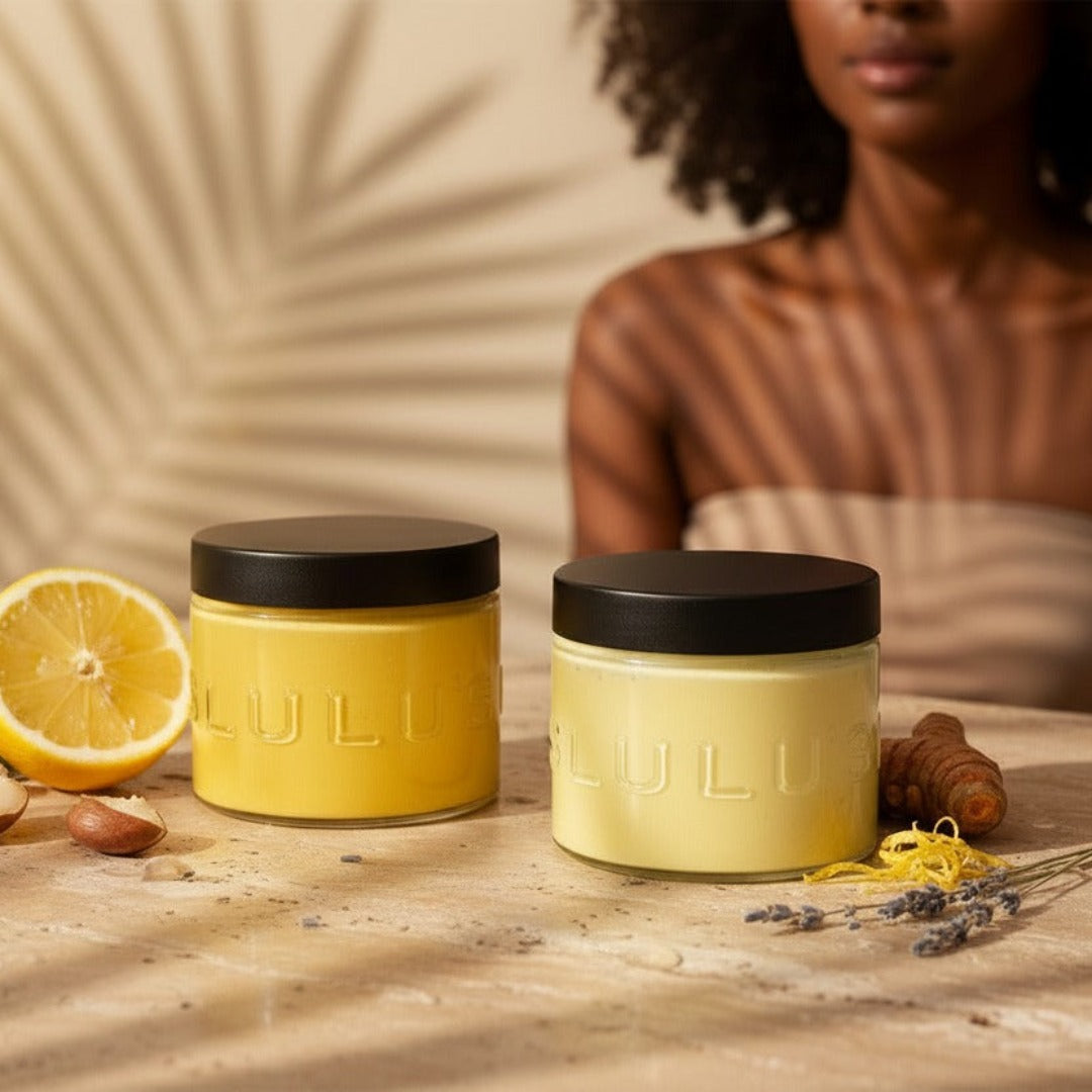 Two jars of Lulu's Holistics Lemon Turmeric Body Butter styled with sliced fresh lemon, raw shea nuts, and turmeric root.