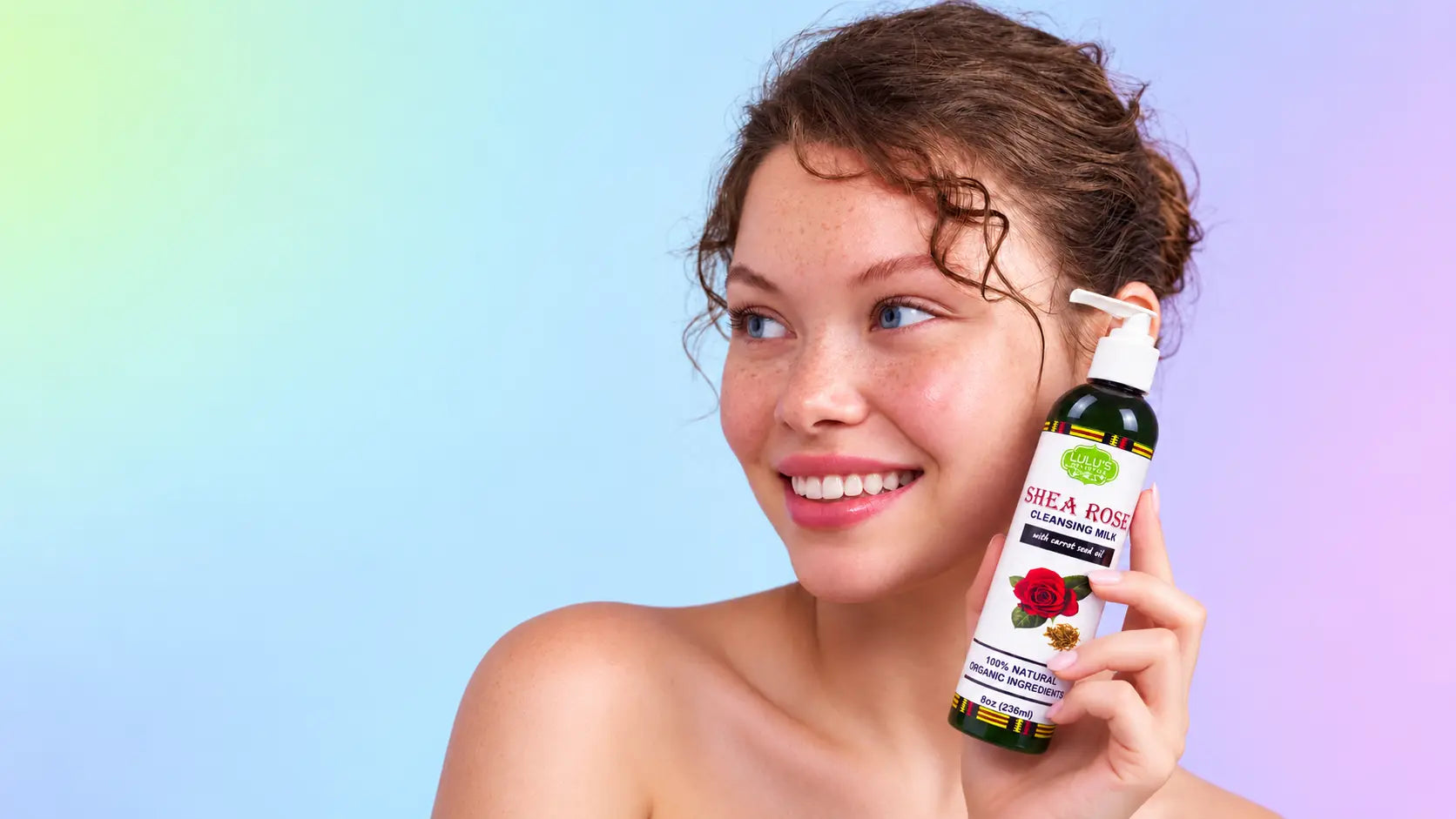 Smiling young woman with natural curly brown hair and visible freckles, holding up a green pump bottle of Lulu's Holistics Shea Rose Cleansing Milk against a soft, pastel rainbow gradient background.