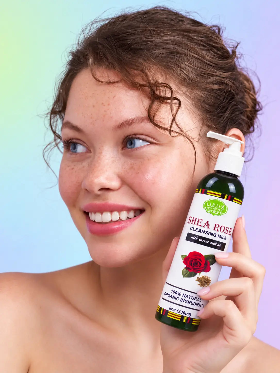 Smiling model with radiant freckled skin holding an eight-ounce bottle of Lulu's Holistics Shea Rose Cleansing Milk with carrot seed oil against a soft pastel gradient background
