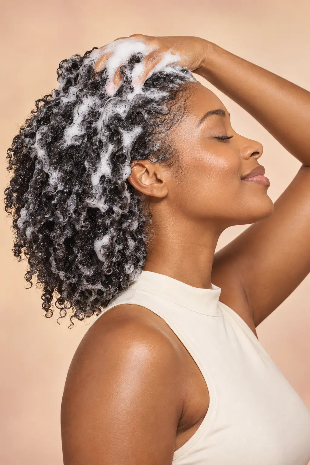 Smiling model with radiant skin massaging rich foamy lather from Lulu's Holistics Vital Castor and Hemp Shampoo into thick defined curls against a tan backdrop